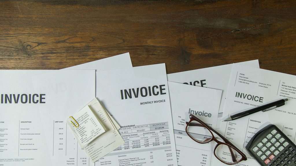 A collection of professional business invoices on a wooden desk with receipts, a pen, glasses, and a calculator for financial management.